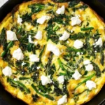 Golden-brown spring vegetable frittata in a cast-iron skillet, topped with crumbled ricotta cheese and fresh greens